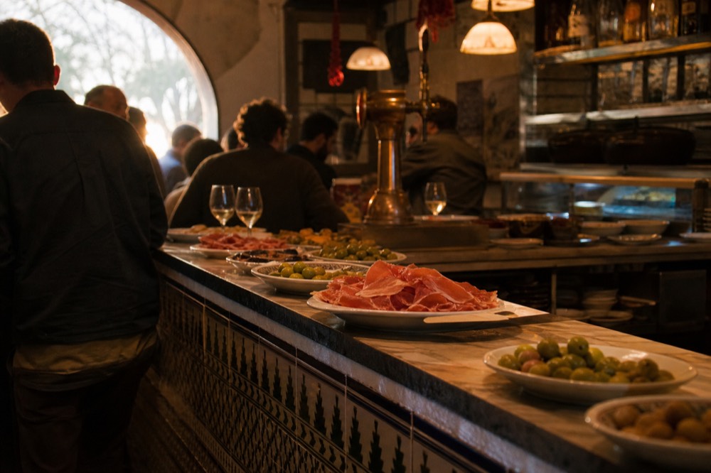 A small tiled tapas bar counter crowded with standing customers and plates of olives and jamon, warm light from overhead lamps