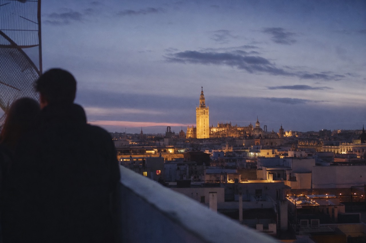 A wide view over Seville rooftops at dusk with the cathedral bell tower lit against a deep blue sky and warm windows below