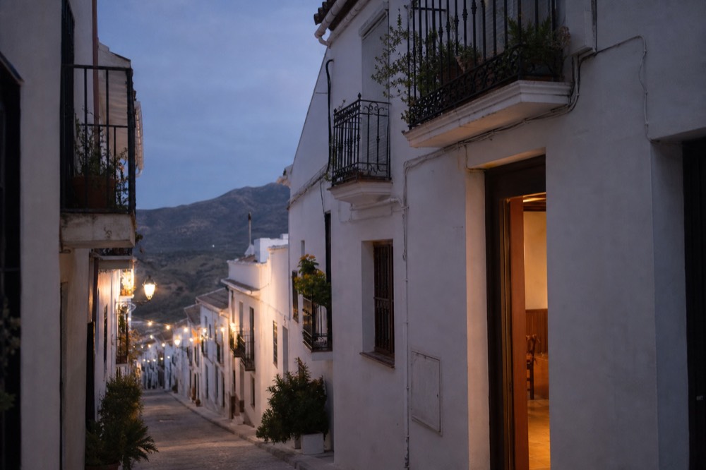 Narrow whitewashed Andalusian street at dusk with iron balconies and one lit doorway, no people in frame