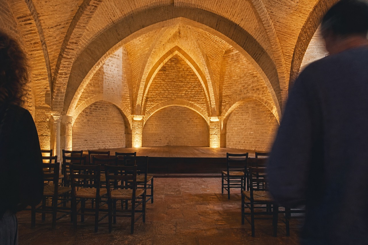 Brick vault with pointed medieval arches lit in amber, two rows of empty chairs facing a small wooden stage