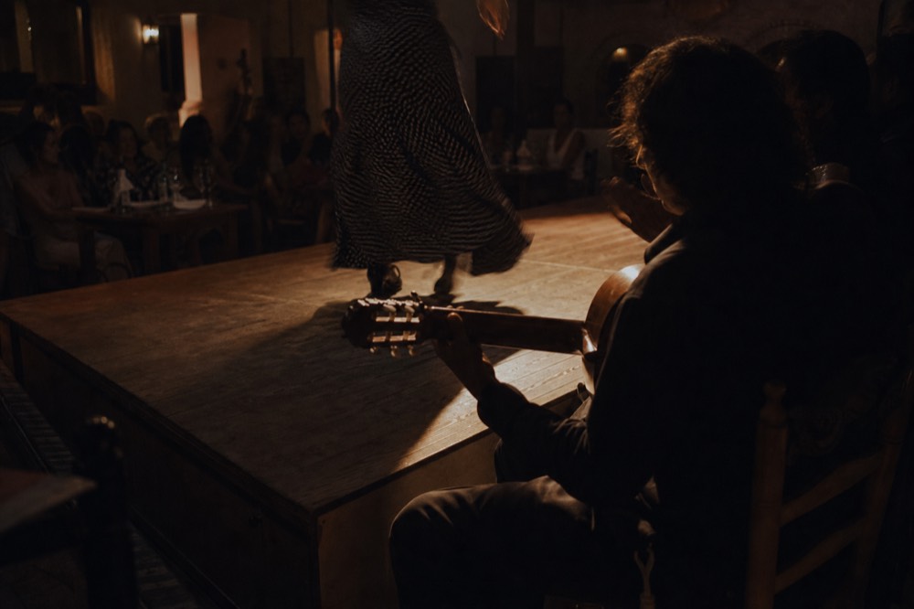 Guitarist and singer seated at the side of a small tablao stage under a single warm spotlight, dancer's feet blurred mid-stamp
