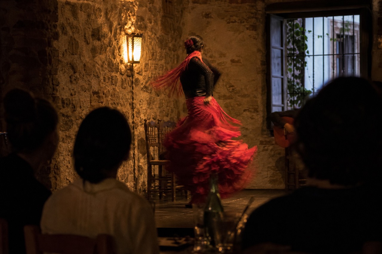 Flamenco dancer mid-turn in a small dark tablao room, skirt blurred with motion, seated audience close to the stage