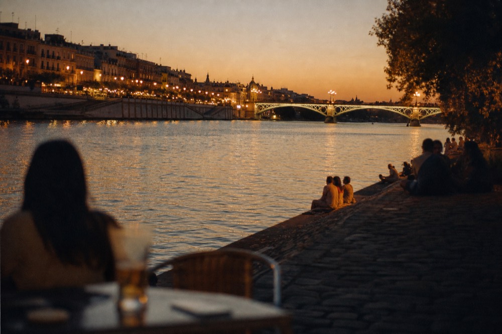 Guadalquivir riverbank at dusk with the Triana bridge lit and lamp-lit bar terraces along the far quay