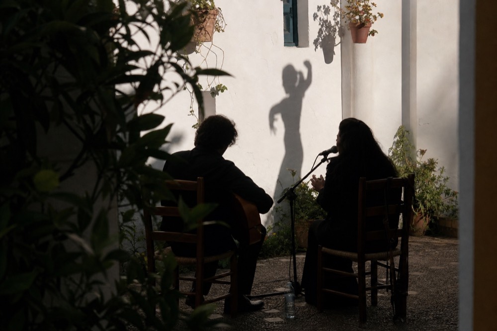 A guitarist and a singer seated on wooden chairs in a small whitewashed courtyard, a dancer's shadow thrown across the wall behind them