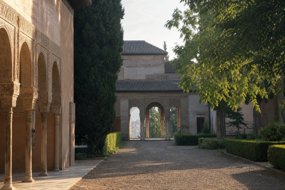 Sunlit Mudejar arches and clipped hedges in a royal palace garden courtyard, empty of people in the early morning