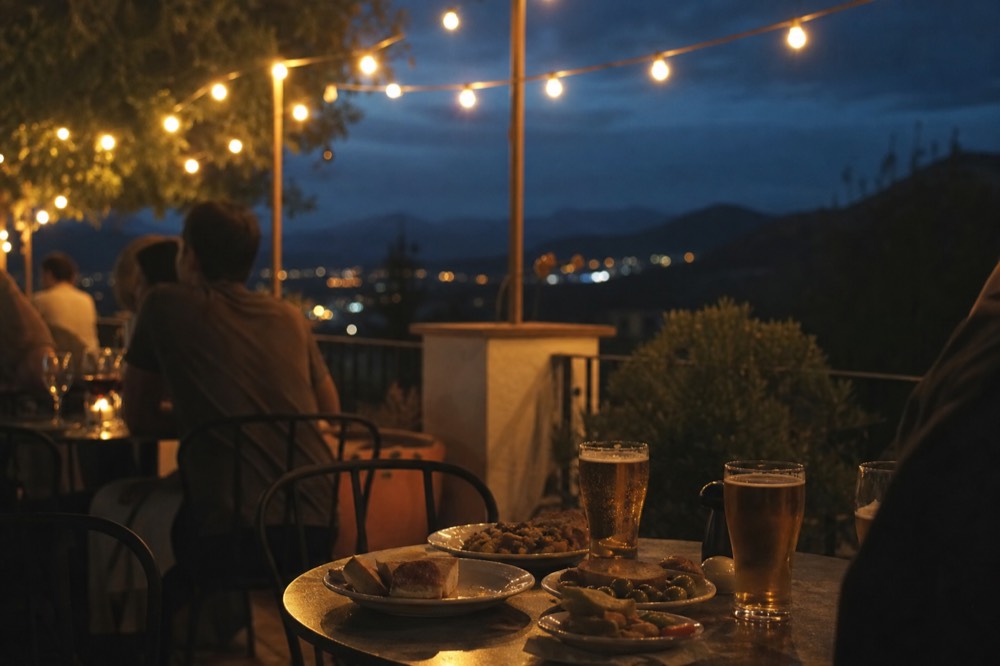 A pavement terrace at night with small round tables, glasses of beer and plates of tapas, people talking under strings of warm bulbs