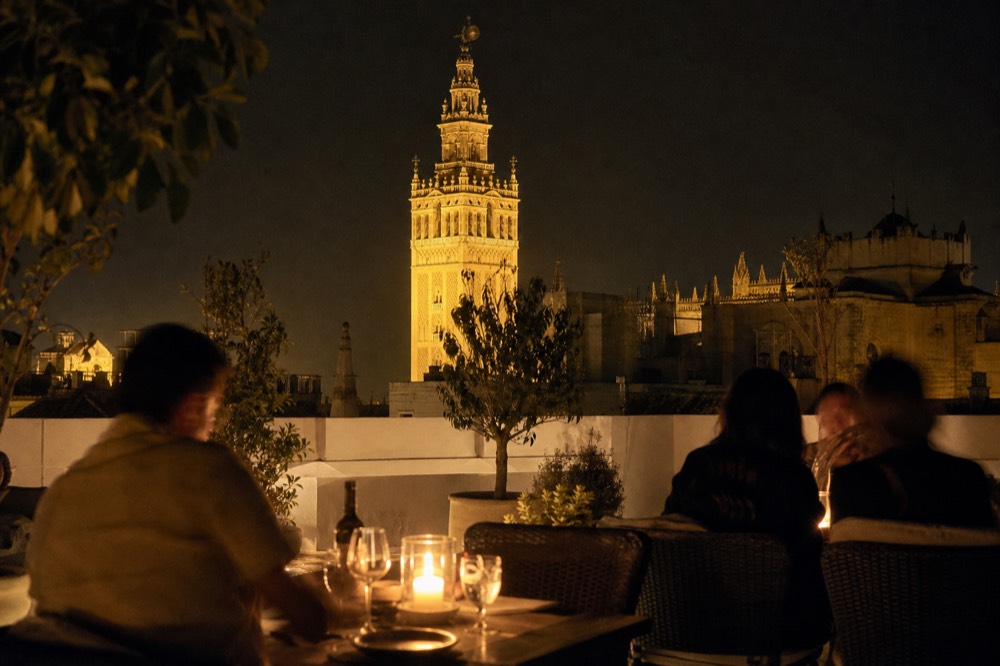 A rooftop terrace at night with candlelit tables and a floodlit cathedral bell tower rising behind the parapet