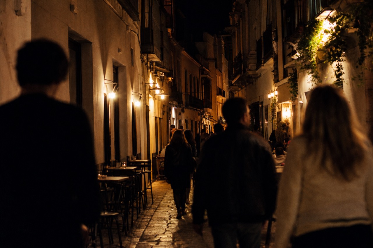 A narrow Seville street after dark lined with lit bar frontages and small pavement tables, a blurred group of people walking between them
