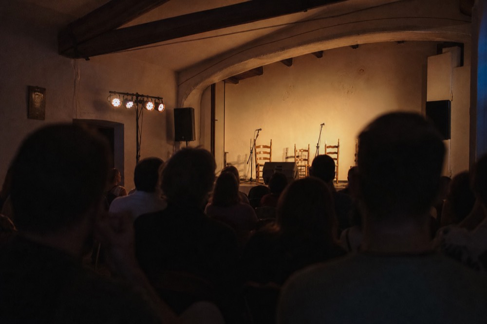 Audience seen from behind in a small dim room, heads and shoulders in silhouette facing a lit empty stage