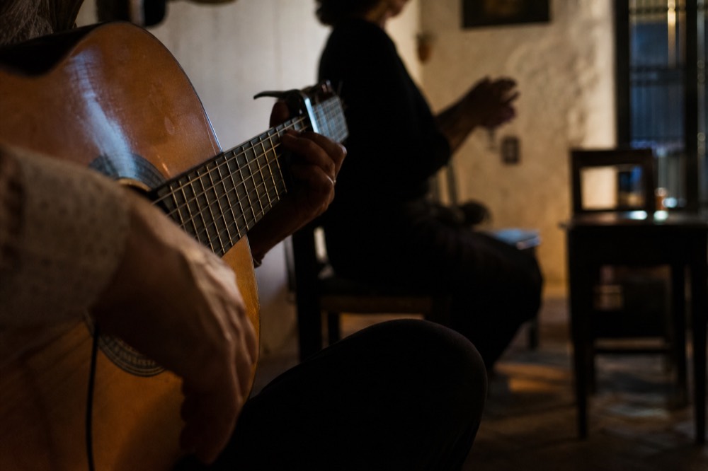 A guitarist's hands close up on the strings with a singer clapping out of focus behind him in a dim room with whitewashed walls