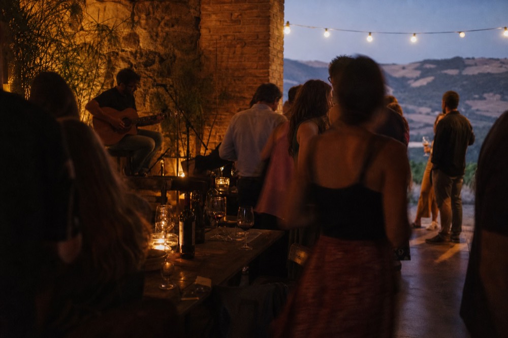 A crowded rustic bar interior with a bare brick wall, a lone guitarist on a stool at one end and drinkers standing between the tables
