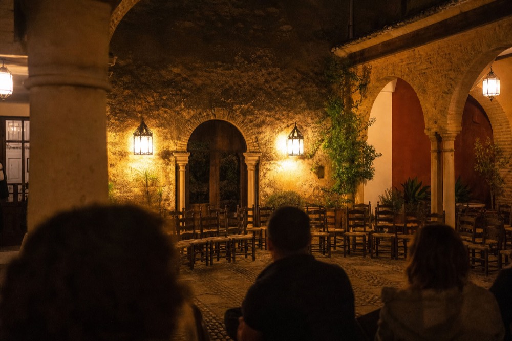 Old Seville courtyard at night with worn stone arches, iron lanterns and empty chairs set out in a semicircle