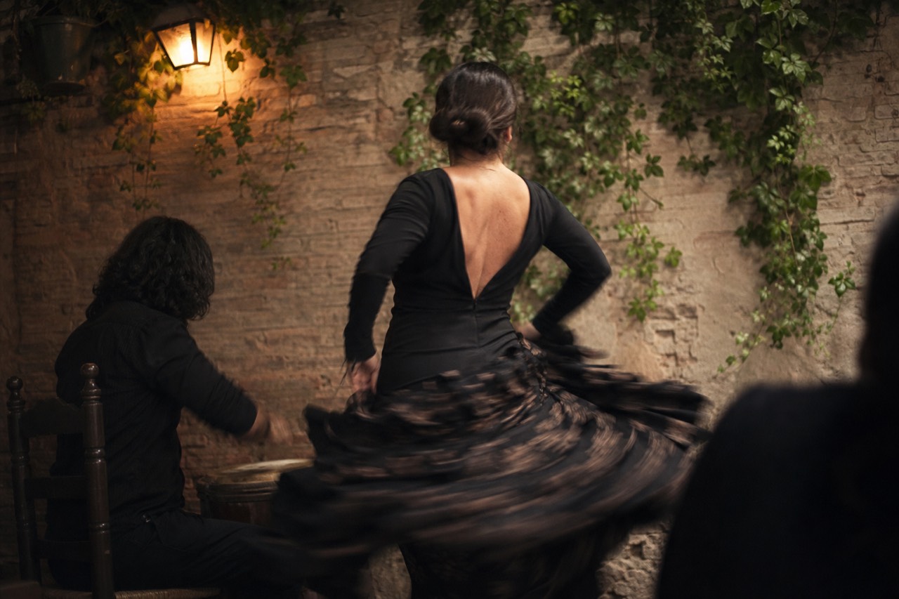 Bare brick tablao wall lit by one warm lamp, a dancer seen from behind mid-turn with her skirt blurred by movement