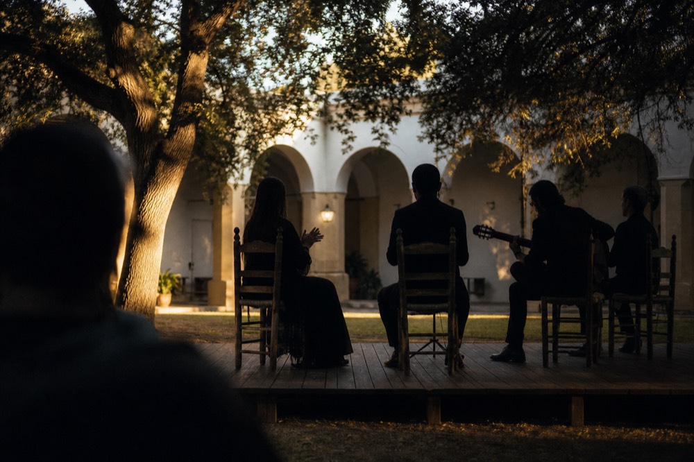 Small unamplified flamenco room in a Seville palace courtyard, four wooden chairs on a low plank stage, whitewashed arches and warm side light