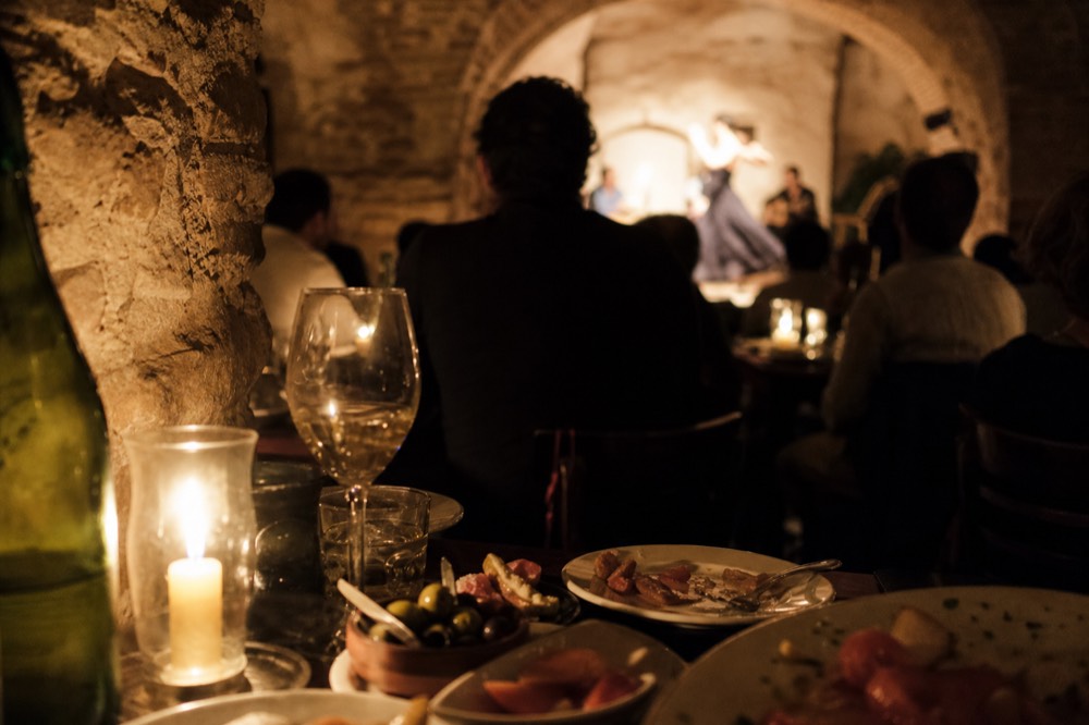 Diners at candlelit tables in a vaulted Seville tablao facing a wooden stage, plates of tapas in the foreground, dancer out of focus beyond