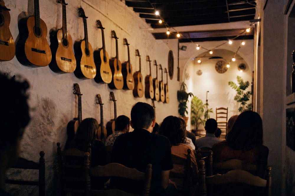 Wall of vintage Spanish guitars hanging in a narrow whitewashed Seville room with a row of wooden chairs facing a low stage