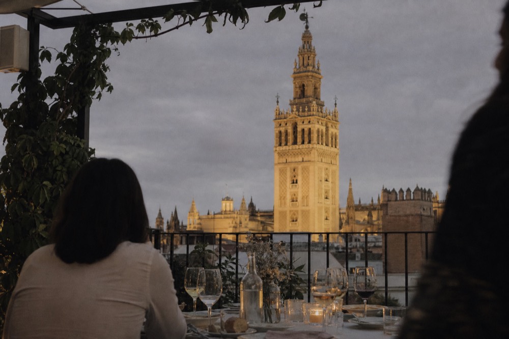 Rooftop dinner table set with wine glasses at dusk in Seville, the illuminated cathedral bell tower rising beyond the terrace railing
