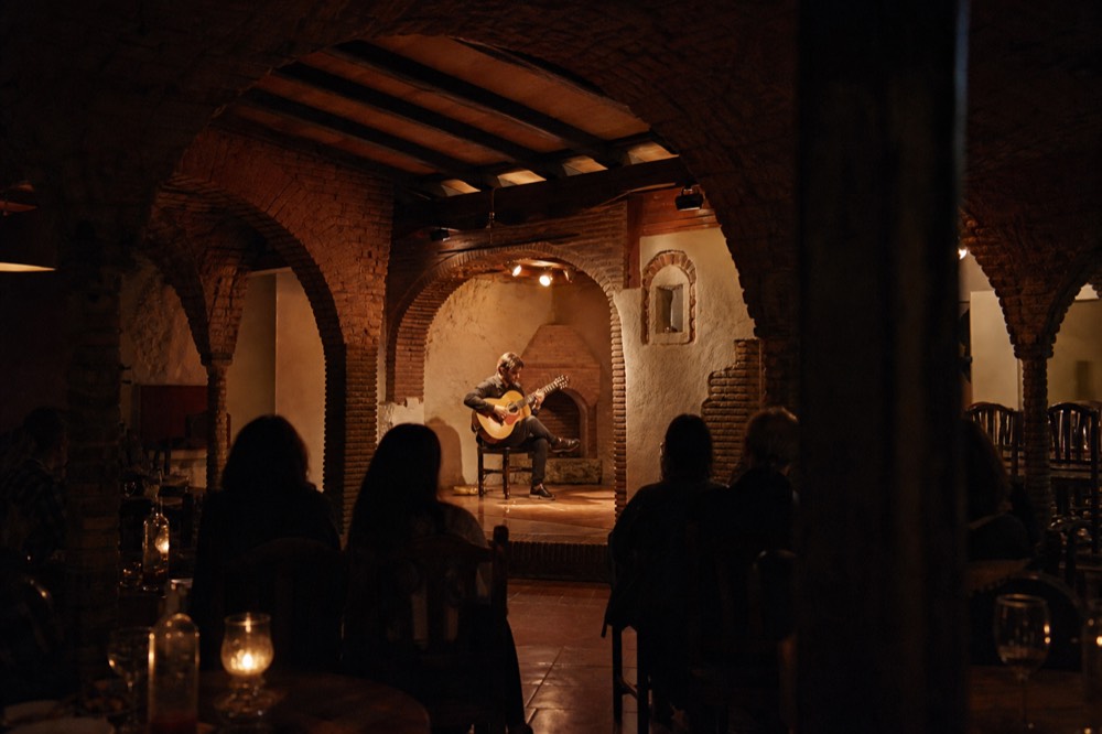 Interior of an old brick-and-timber tablao room with dinner tables ringing a low stage, a guitarist seated in warm light