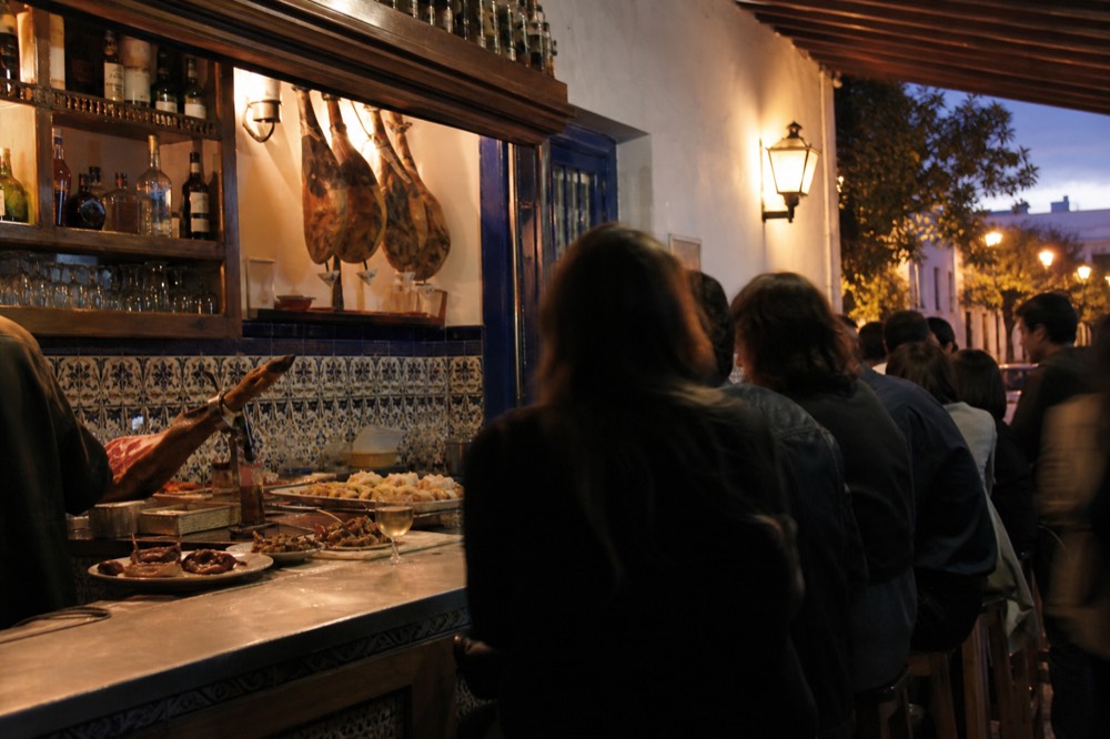 Crowded tapas bar counter in Triana with tiled walls, hanging hams and small plates lined up along the zinc top