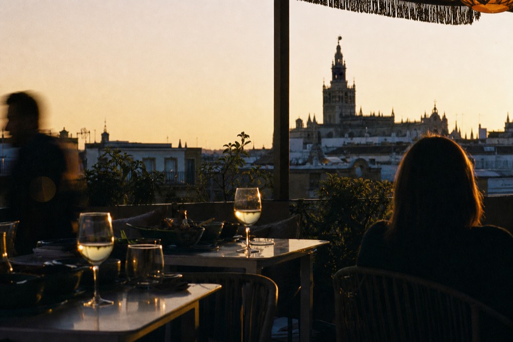 Rooftop terrace tables at dusk with the silhouette of the Seville cathedral tower behind, wine glasses catching the last light