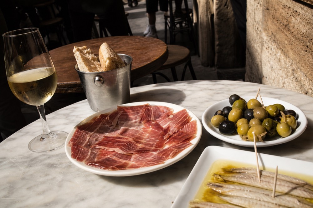 Close view of a tapas spread on a small marble table in a Seville bar, jamon, olives and a glass of chilled sherry