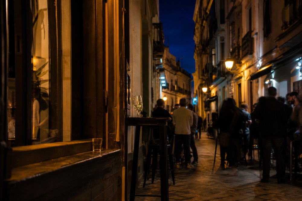 Narrow Seville street after dark with lit bar doorways and people standing at outdoor tables late in the evening