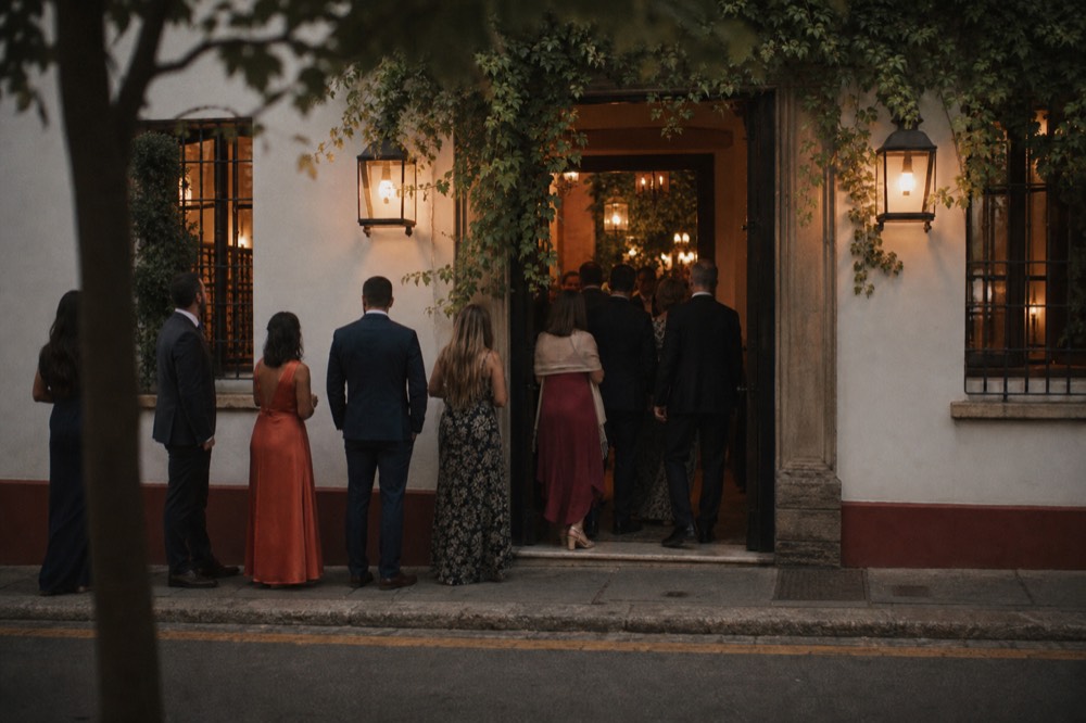 Queue of well-dressed guests waiting at a lantern-lit venue doorway on a warm Seville evening, seen from across the street