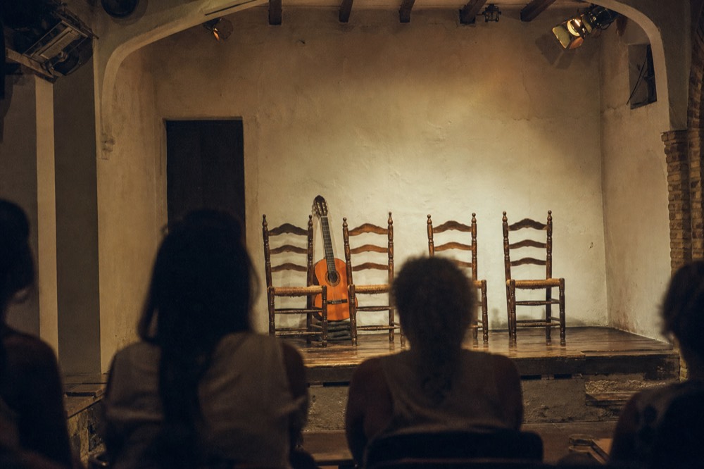 Four empty chairs and a bare wooden stage in a small flamenco room, single guitar resting against a chair, warm spotlights off