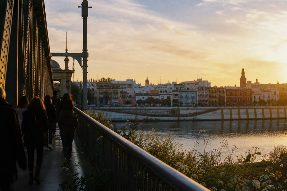 View along an old iron bridge over the Guadalquivir at golden hour, tiled Triana rooftops on the far bank, pedestrians in silhouette