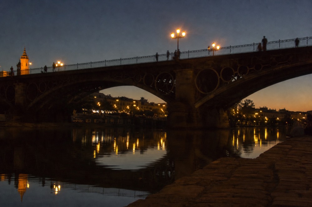 The old iron bridge to Triana photographed at night from the riverbank, lamps reflected in still water, silhouetted walkers on the span