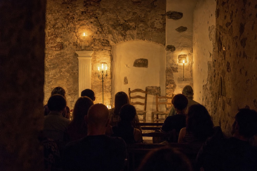 Small audience seated two rows deep in a candlelit stone room waiting for a flamenco performance to begin