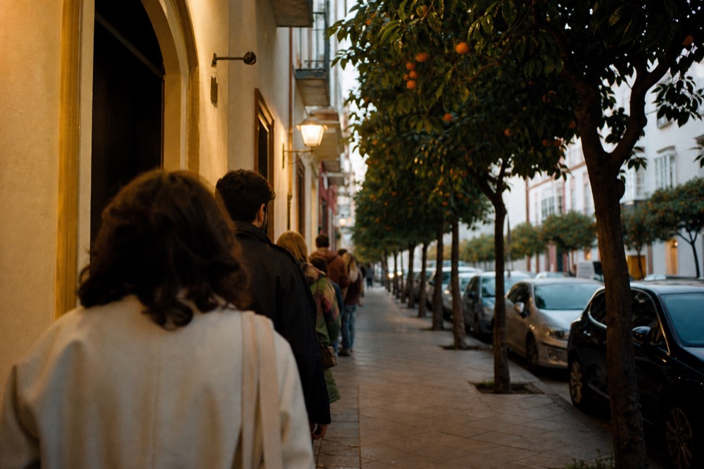 Seville street in late afternoon light with orange trees and a queue forming outside a small venue door
