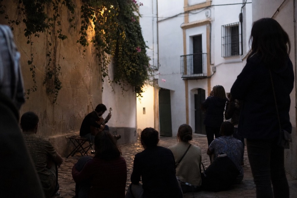 Small crowd gathered loosely around a guitarist seated on a folding stool in a Seville street at dusk, faces turned away from the camera