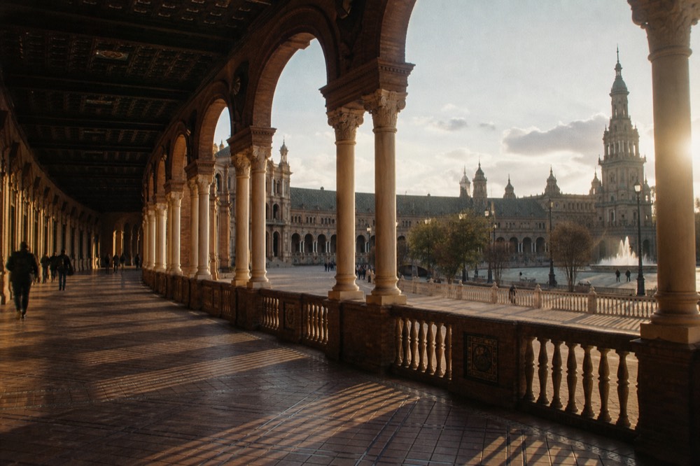 Wide tiled colonnade of a grand Seville plaza in late afternoon, long shadows across the paving, scattered visitors in the distance