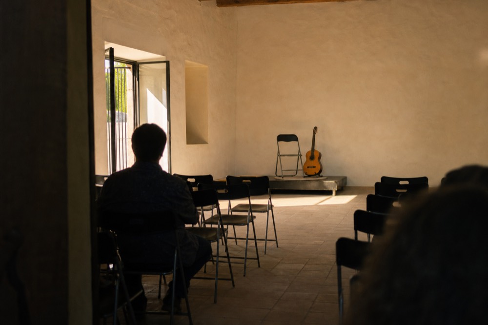 Plain club room in Andalusia with folding chairs in rows, a single chair and guitar on a low platform, bare walls and a tiled floor