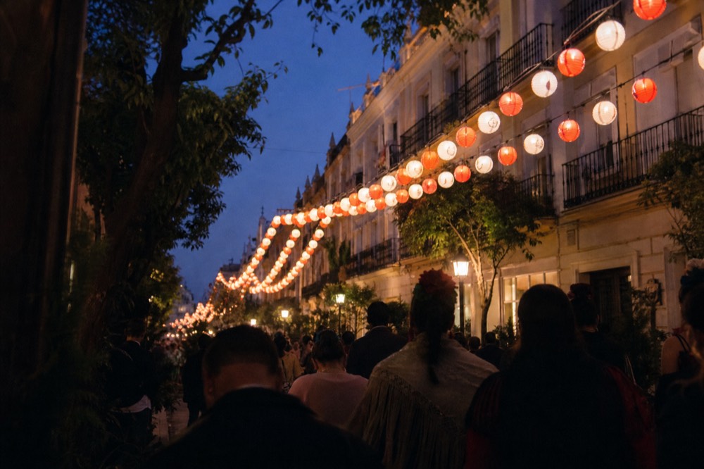 Crowded Seville street during a spring festival evening, strings of lanterns overhead, people in silhouette against warm light