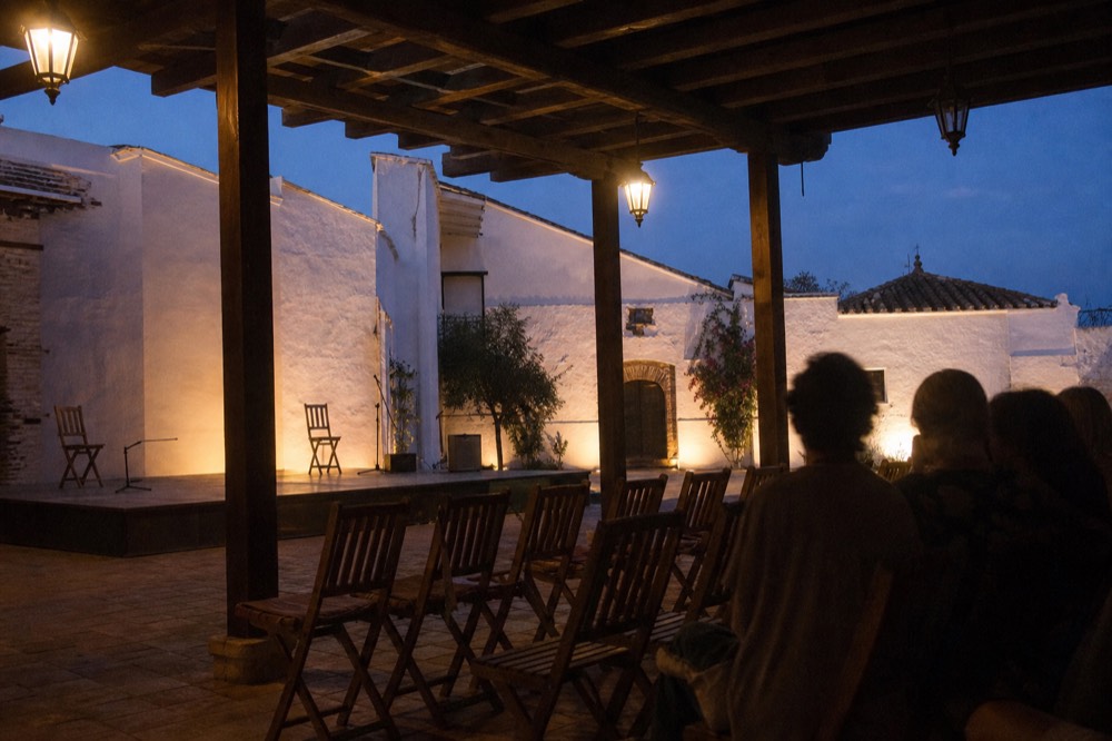 Two rows of empty wooden chairs facing a small raised stage in a whitewashed Andalusian courtyard, evening light on the upper walls