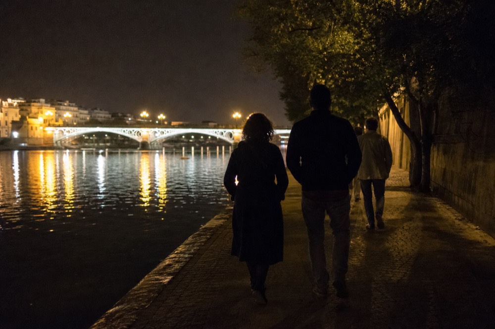 Riverside walkway in Seville after dark, bridge lights reflected in the water, a few people walking away from the camera