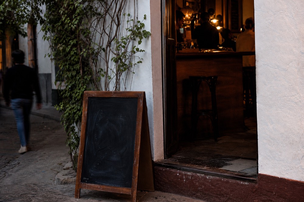 Hand-lettered chalkboard propped by a bar doorway in Seville at dusk, no readable text, warm light spilling onto cobbles
