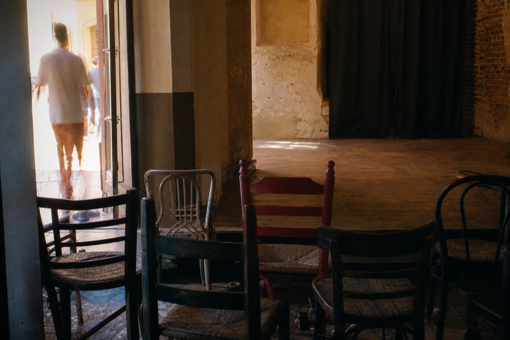 Empty mismatched chairs facing a bare wooden performance floor in a Seville bar in the last hour before opening, low sun through a doorway