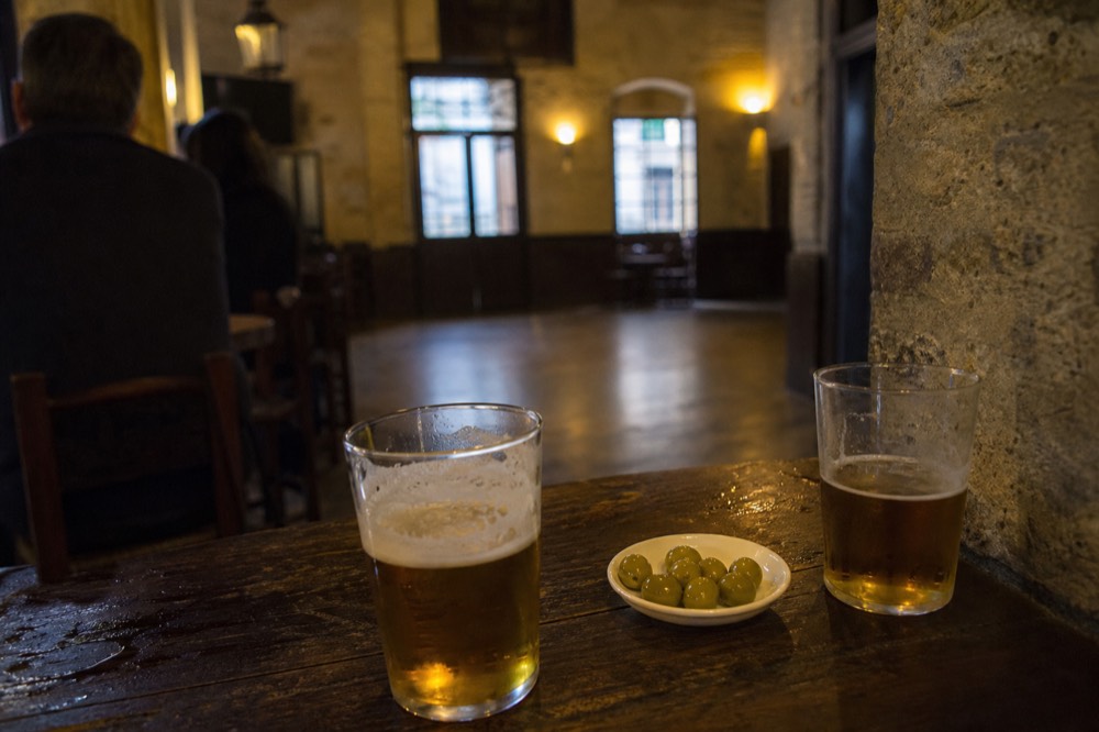 Two glasses of beer and a small plate of olives on a battered wooden table in a Seville bar, dance floor empty in the soft-focus background
