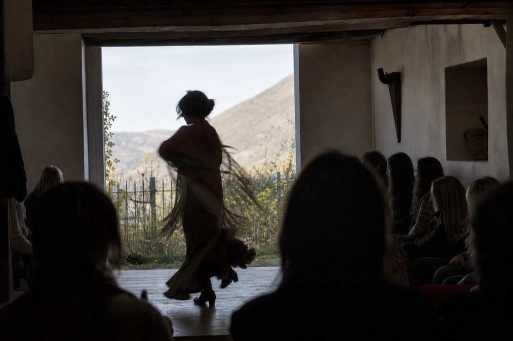 Silhouette of a flamenco dancer mid-turn on a small wooden floor, seated audience close on three sides, shot from behind the last row