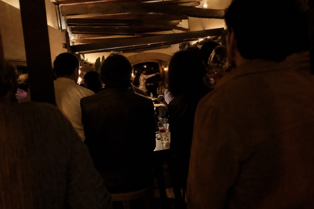 Crowded Seville bar interior at night seen over the heads of standing drinkers, small lit performance floor barely visible at the far end