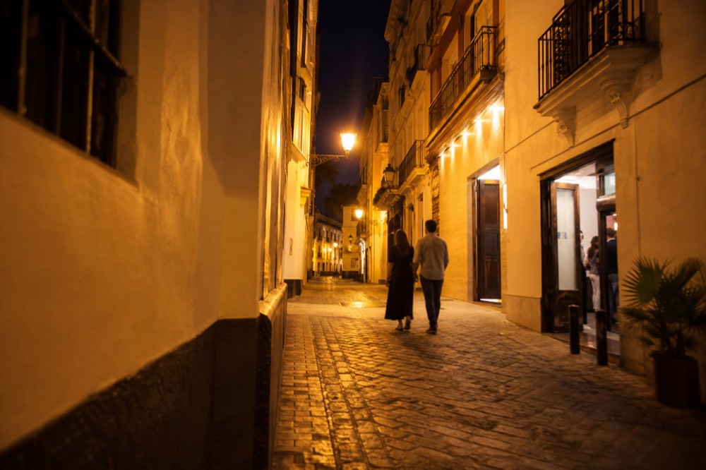Narrow Seville street at night with orange street lamps, a couple walking away from camera toward a lit bar doorway