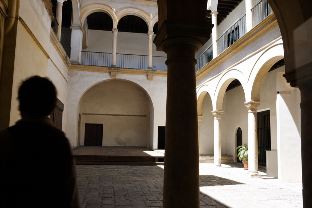 Interior of an old Seville townhouse with a stone-flagged patio, arched galleries on two levels and a bare wooden stage at one end