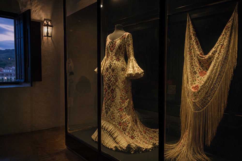 Glass display case of embroidered flamenco costume and a fringed shawl in a dimly lit museum room, no visitors in frame