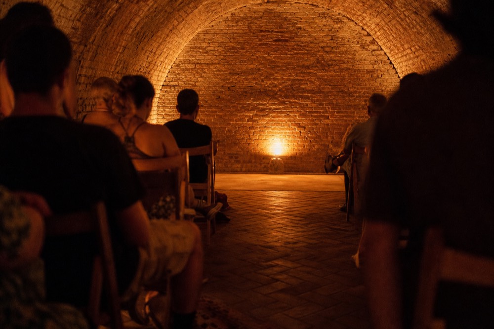 Brick vaulted cellar with two short rows of wooden chairs facing a small clear floor, single warm lamp low on the wall