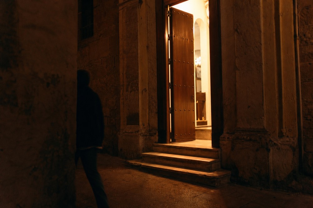 Old Seville street doorway at night with a heavy wooden door ajar and warm light spilling out onto worn stone steps