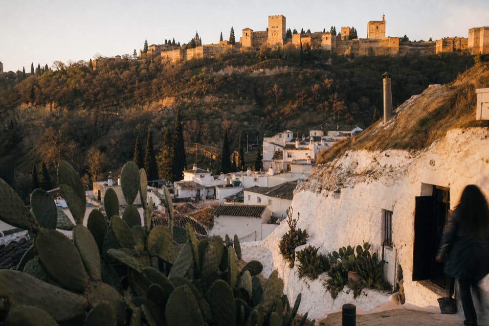 Whitewashed cave dwellings on the Sacromonte hillside at dusk with prickly pear in the foreground and the Alhambra walls beyond