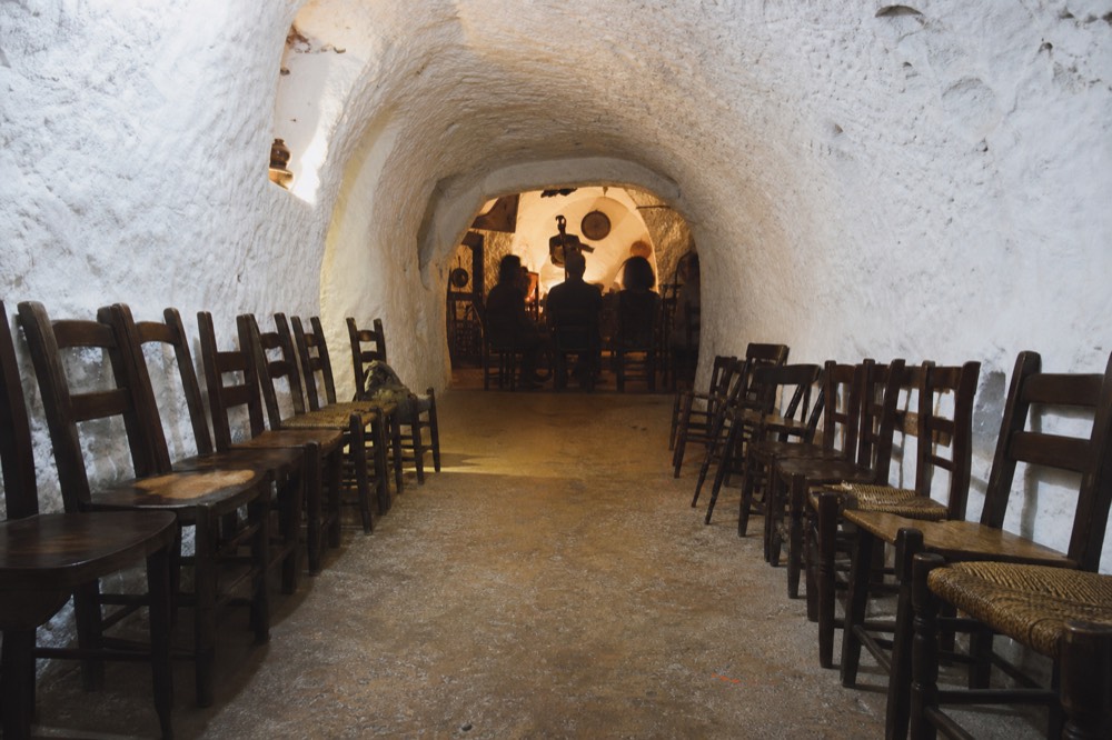 Interior of a narrow whitewashed cave venue with vaulted ceiling, wooden chairs lined along both side walls and a bare floor between them